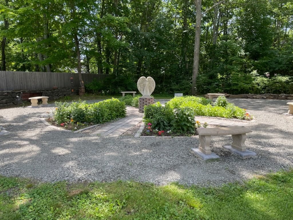 The All Saints' Prayer and Meditation Garden with stone benches, flowering beds, and an angel sculpture beneath shade trees
